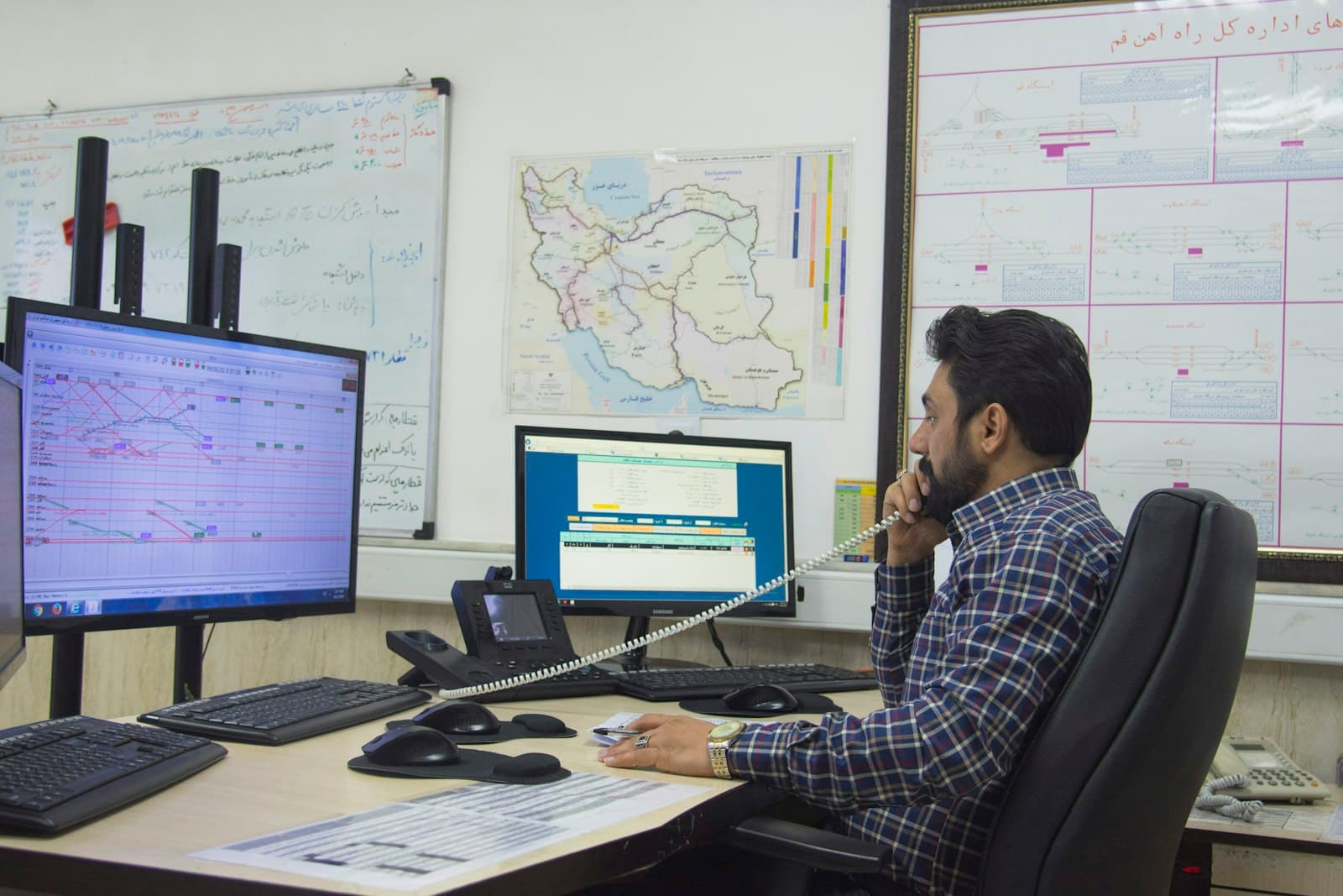 A dispatcher on the phone at a desk with route maps and two monitors